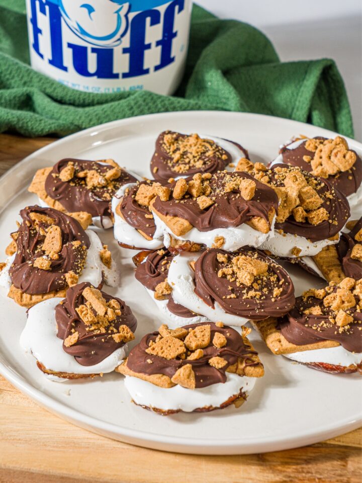 A bunch of smashed date s'mores on a white plate that is on a wooden cutting board. Behind the plate is a green kitchen towel with a tub of marshmallow fluff on it.