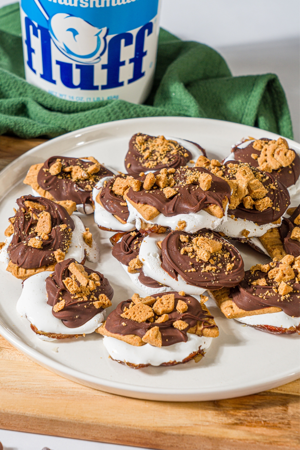 A bunch of smashed date s'mores on a white plate that is on a wooden cutting board. Behind the plate is a green kitchen towel with a tub of marshmallow fluff on it.