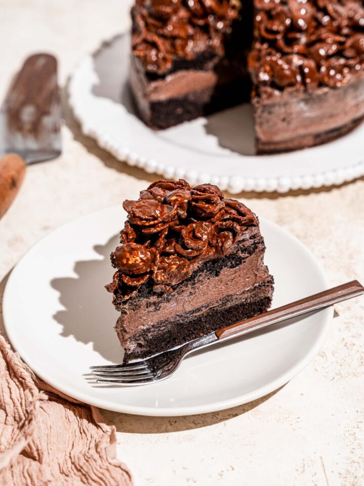 A white plate with a slice of chocolate crunch cake with a fork on the plate. The plate is on a tan counter with a white platter of cake.