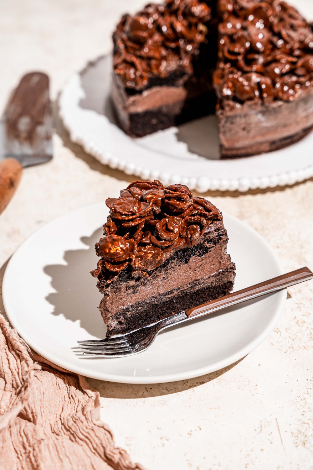 A white plate with a slice of chocolate crunch cake with a fork on the plate. The plate is on a tan counter with a white platter of cake.