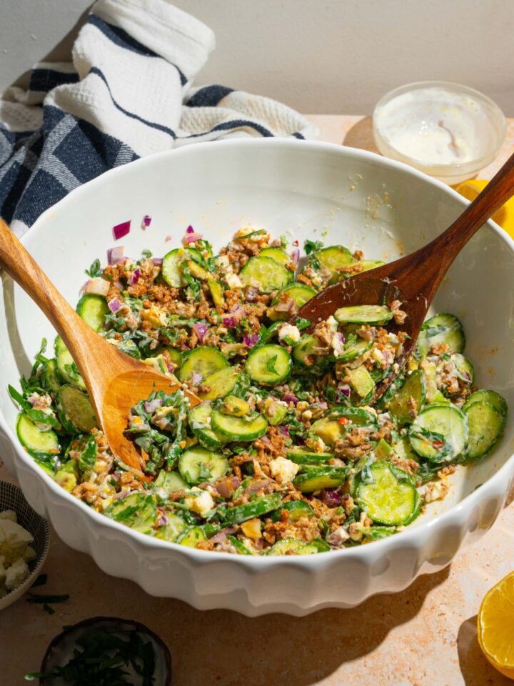 A large bowl with high protein cucumber salad ingredients piled with two salad tongs. The bowl contains cucumbers, feta cheese, avocado, seasoned ground beef, red onion, and greek yogurt dressing. The bowl is on a tan counter.