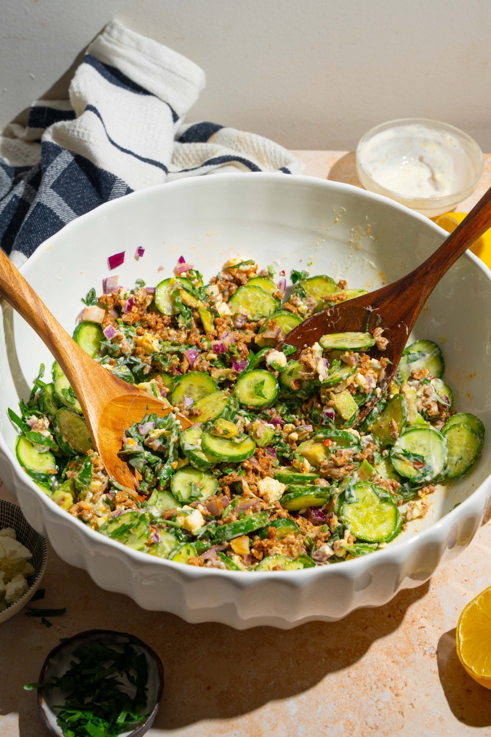 A large bowl with high protein cucumber salad ingredients piled with two salad tongs. The bowl contains cucumbers, feta cheese, avocado, seasoned ground beef, red onion, and greek yogurt dressing. The bowl is on a tan counter.