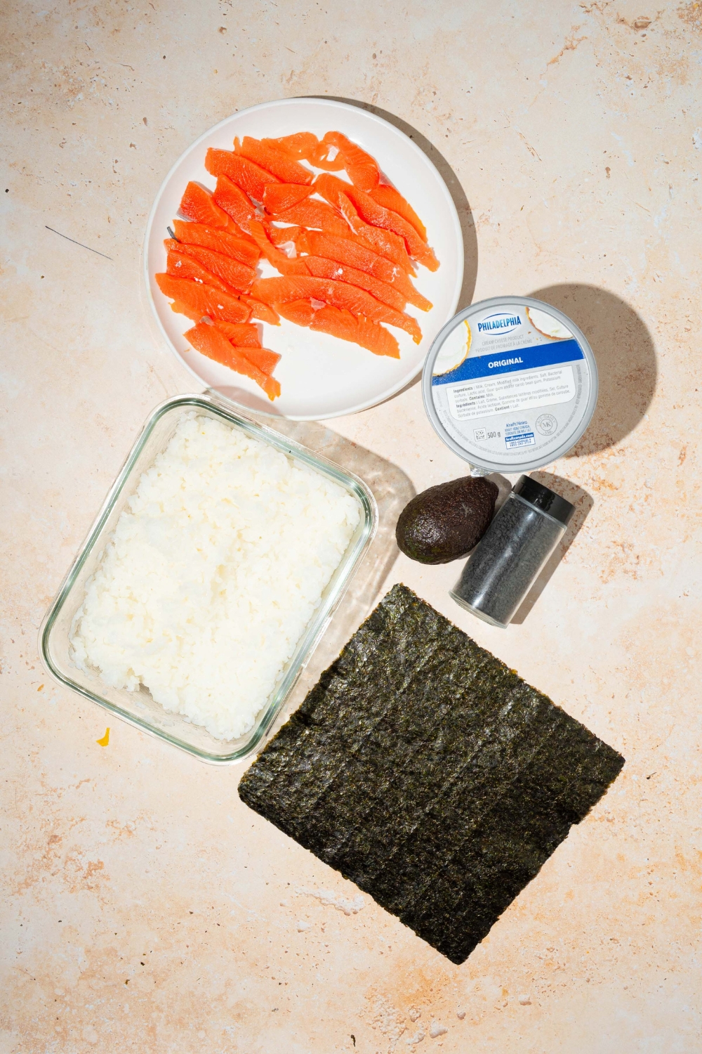 A tan counter with ingredients to make lazy sushi including smoked salmon, cream cheese, nori, rice, avocado, and sesame seeds.