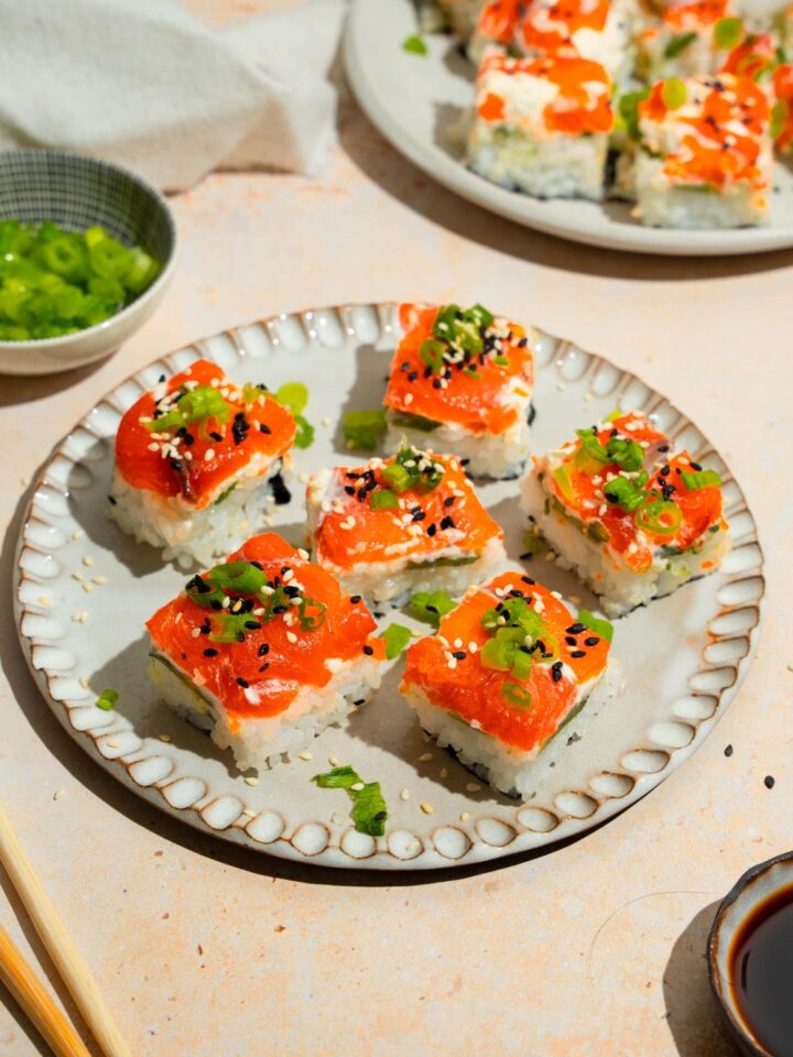 A plate of lazy sushi with salmon topped with sliced green onion and sesame seeds. The plate is on a tan counter with a plate of soy sauce and an additional plate of sushi.