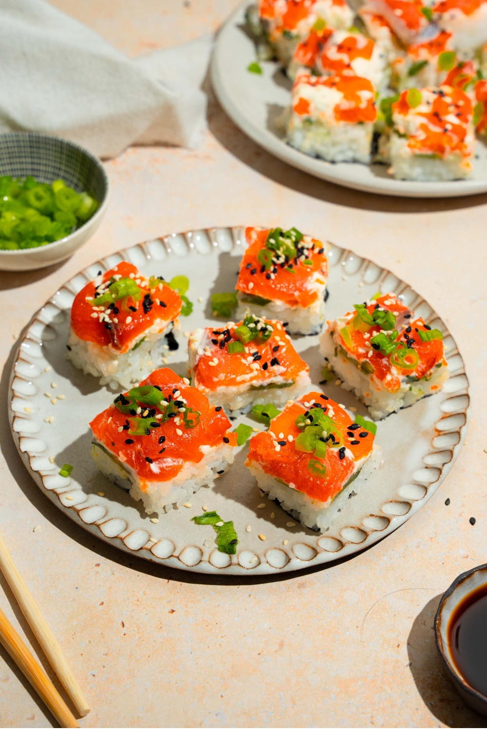 A plate of lazy sushi with salmon topped with sliced green onion and sesame seeds. The plate is on a tan counter with a plate of soy sauce and an additional plate of sushi.