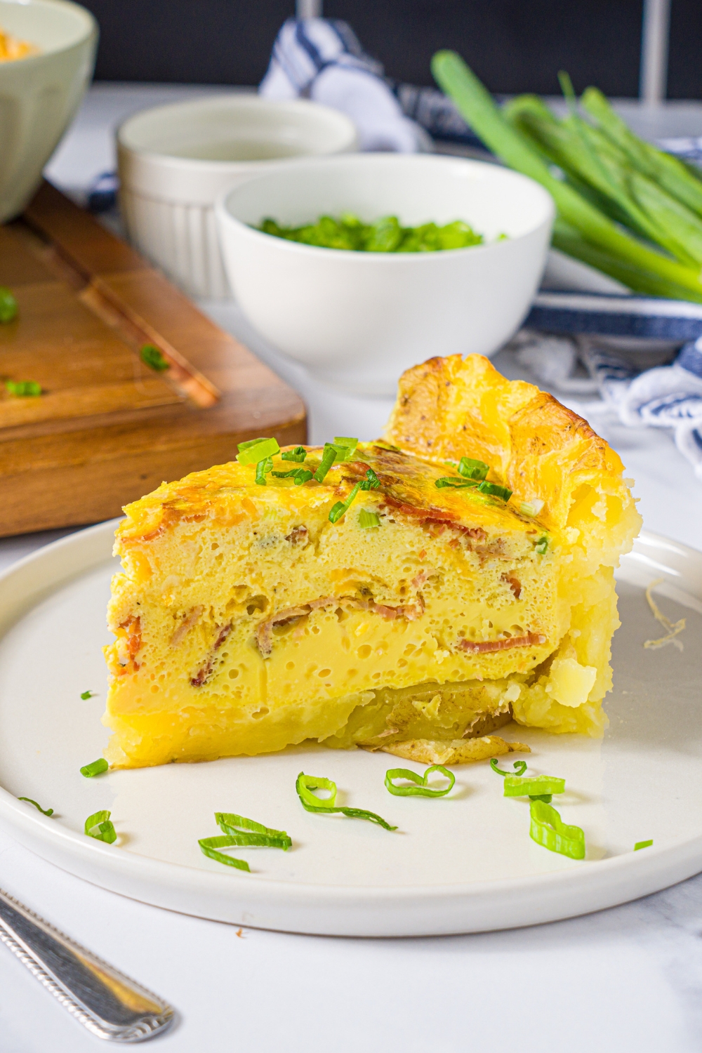 A white plate with a slice of potato crust quiche garnished with sliced green onion. The plate is on a marble counter with a bowl of garnishes.