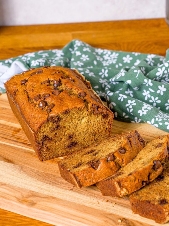 A loaf of banana bread with oil on a wooden cutting board with three slices overlapping one another in front of the loaf.