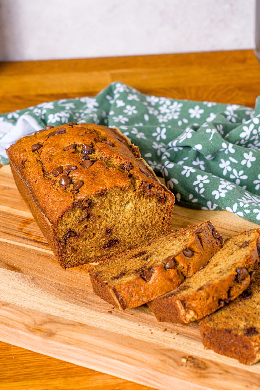A loaf of banana bread with oil on a wooden cutting board with three slices overlapping one another in front of the loaf.
