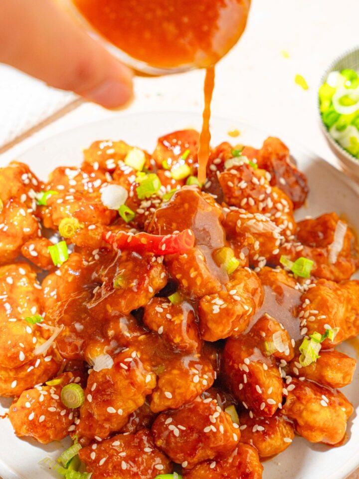 A white plate with crispy general tso's chicken with a jar of sauce being poured over the chicken. The plate is garnished with sliced green onion, sesame seeds, and red chile.