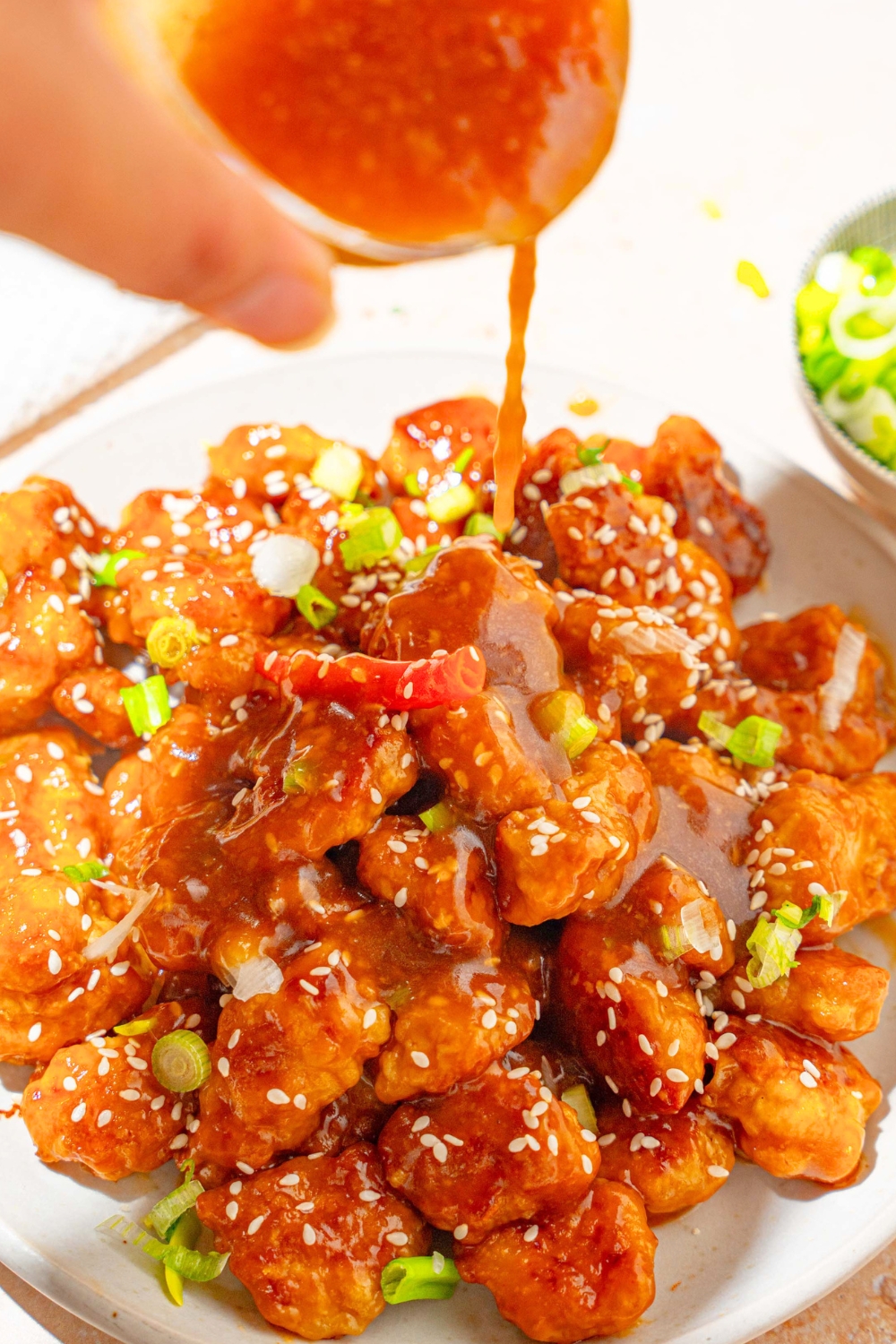 A white plate with crispy general tso's chicken with a jar of sauce being poured over the chicken. The plate is garnished with sliced green onion, sesame seeds, and red chile.