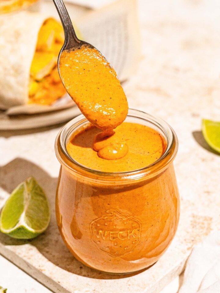 A spoon covered in burrito sauce drizzling it into a glass jar that is filled with the burrito sauce.