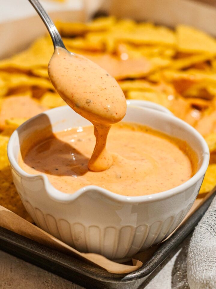 A spoon with cheese sauce on it and pouring down for it into a white bowl filled with the cheese sauce.