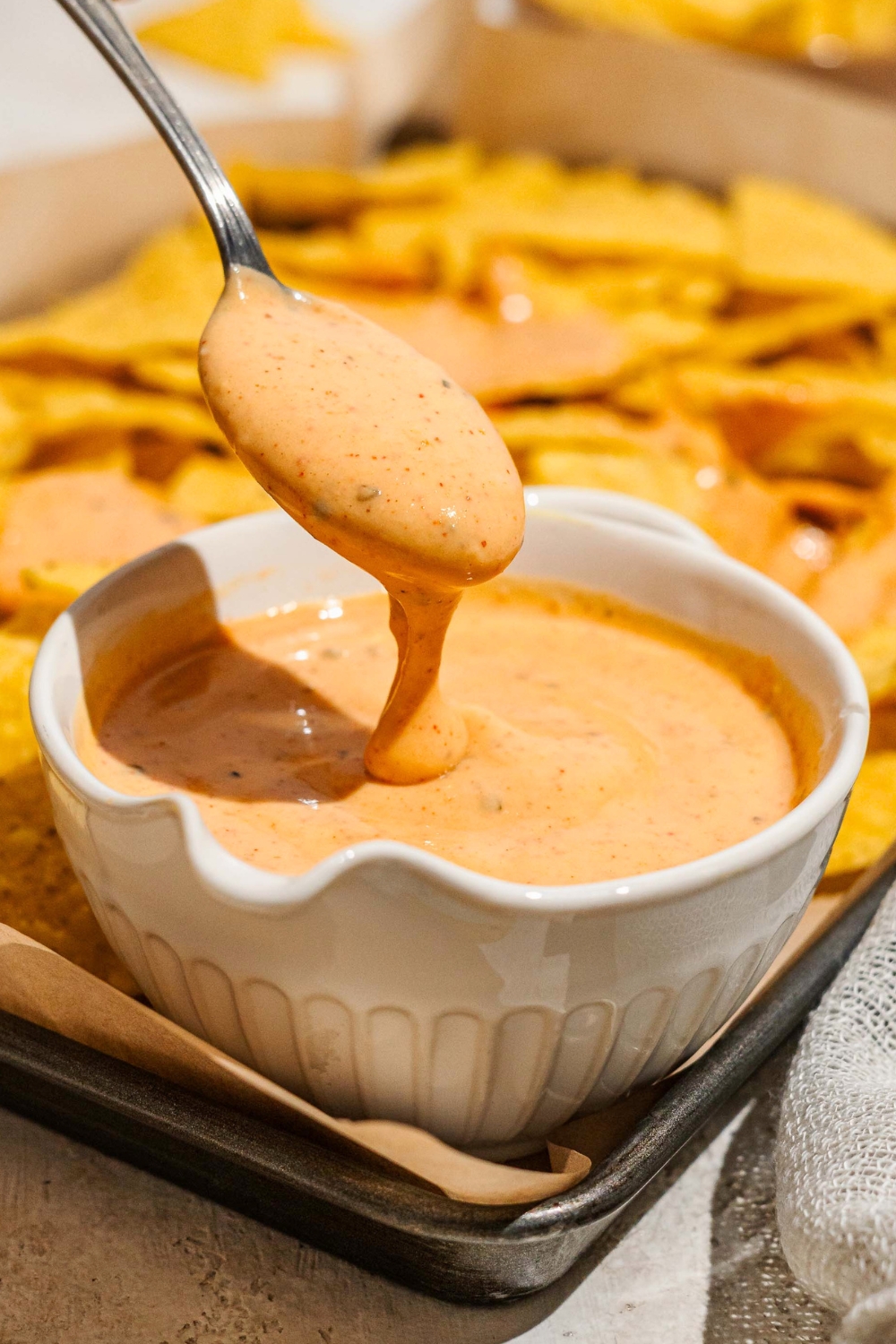 A spoon with cheese sauce on it and pouring down for it into a white bowl filled with the cheese sauce.