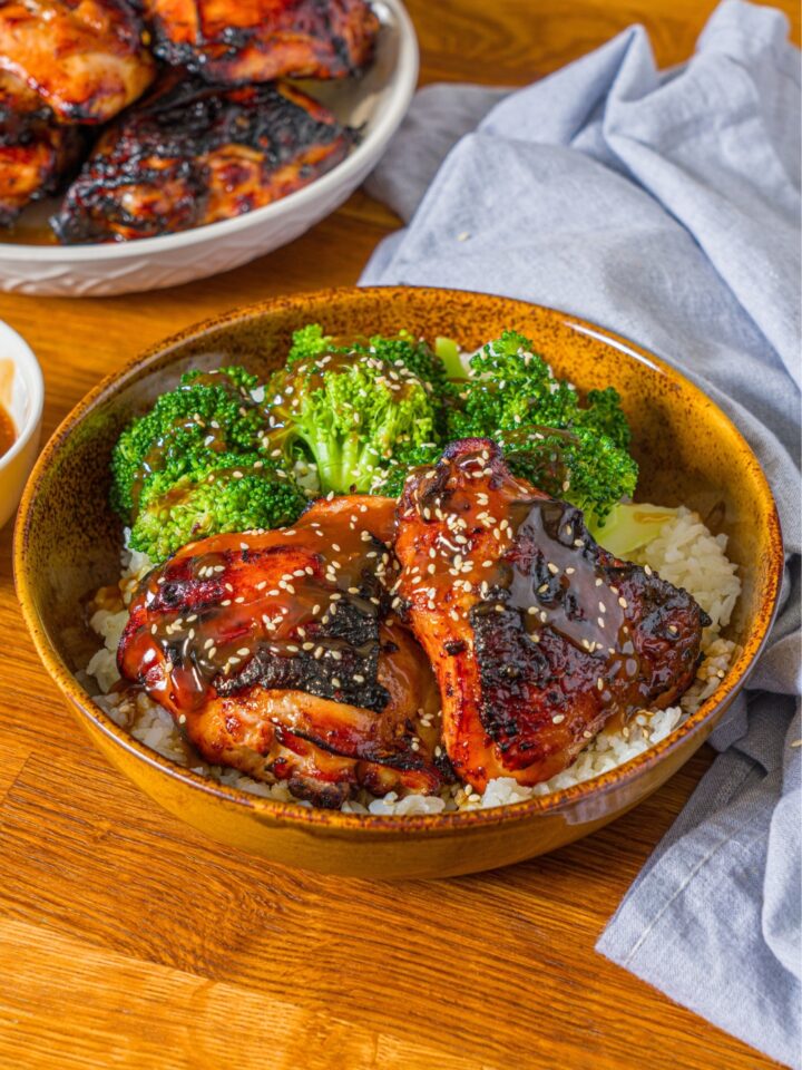 A chicken teriyaki bowl with two chicken teriyaki thighs coated in teriyaki sauce on a bed of basmati rice with broccoli in a wooden bowl.