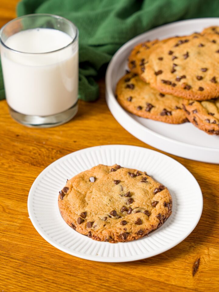 A white plate with a single chocolate chip cookie without baking soda. The plate is on a wooden counter with an additional plate of cookies and a glass of milk.