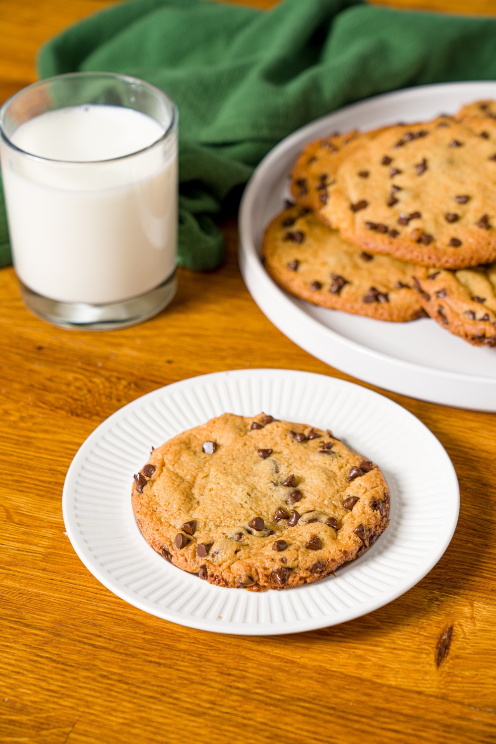 A white plate with a single chocolate chip cookie without baking soda. The plate is on a wooden counter with an additional plate of cookies and a glass of milk.
