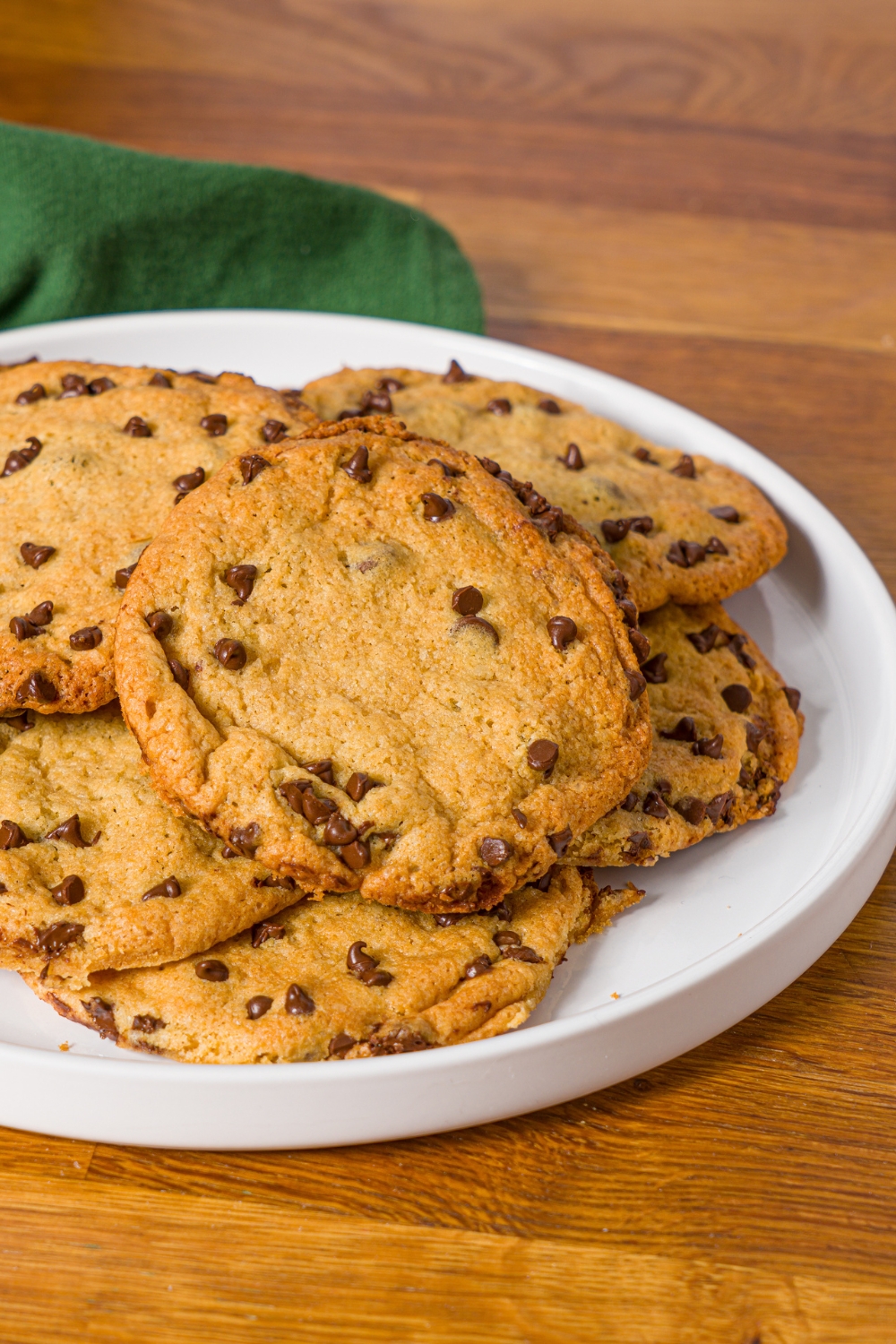 A white plate with chocolate chip cookies without baking soda. The plate is on a wooden counter with a green cloth napkin.