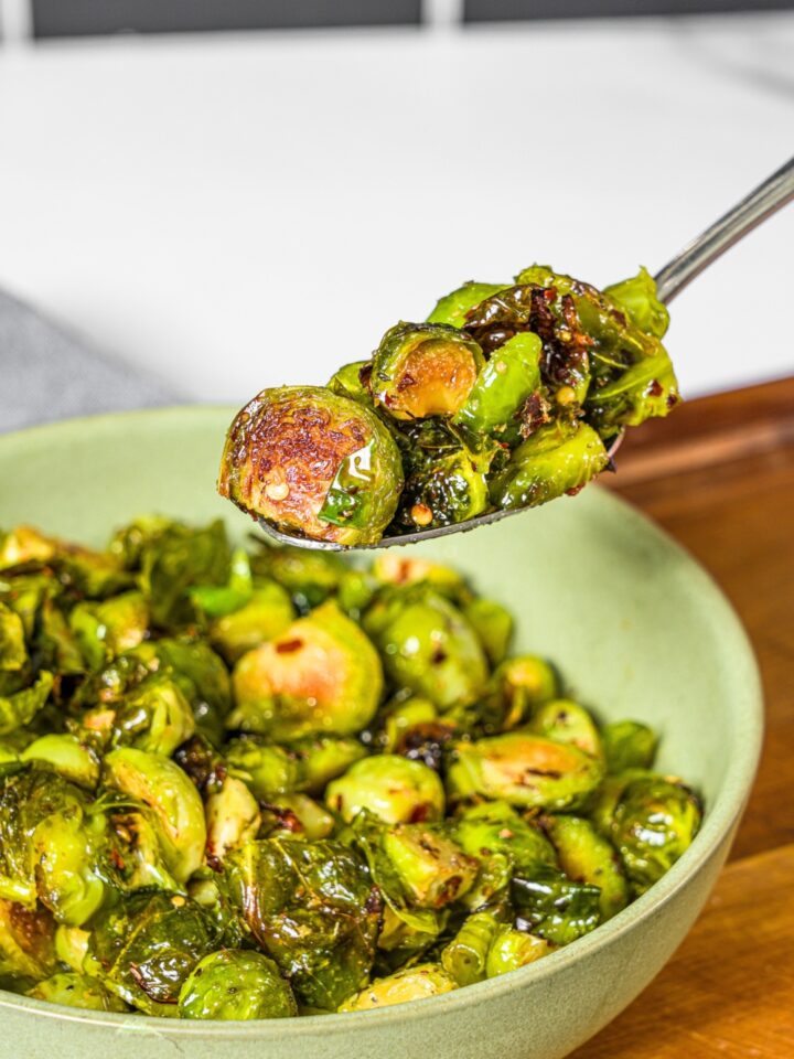 A spoon with crispy brussel sprouts on it being held above a bowl filled with the crispy brussel sprouts.