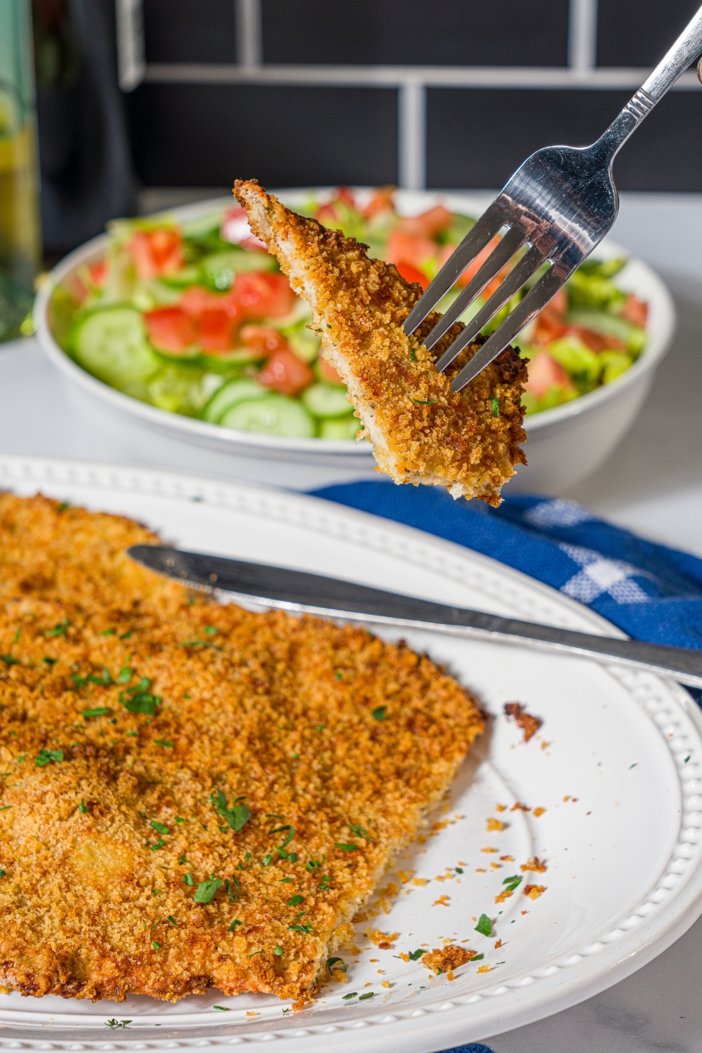 With a piece of a ground chicken cutlet on it above a white plate that has the crispy ground chicken cutlet on it.