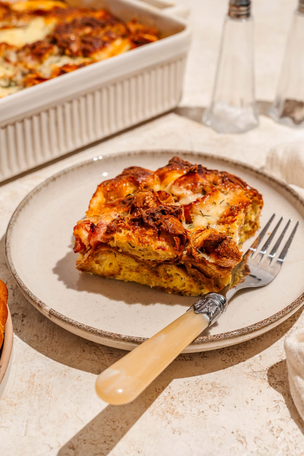 A white plate with a slice of croissant breakfast casserole with ham and cheese. There is a fork on the plate. The plate is on a tan counter with a baking dish of casserole and a salt and pepper shaker.