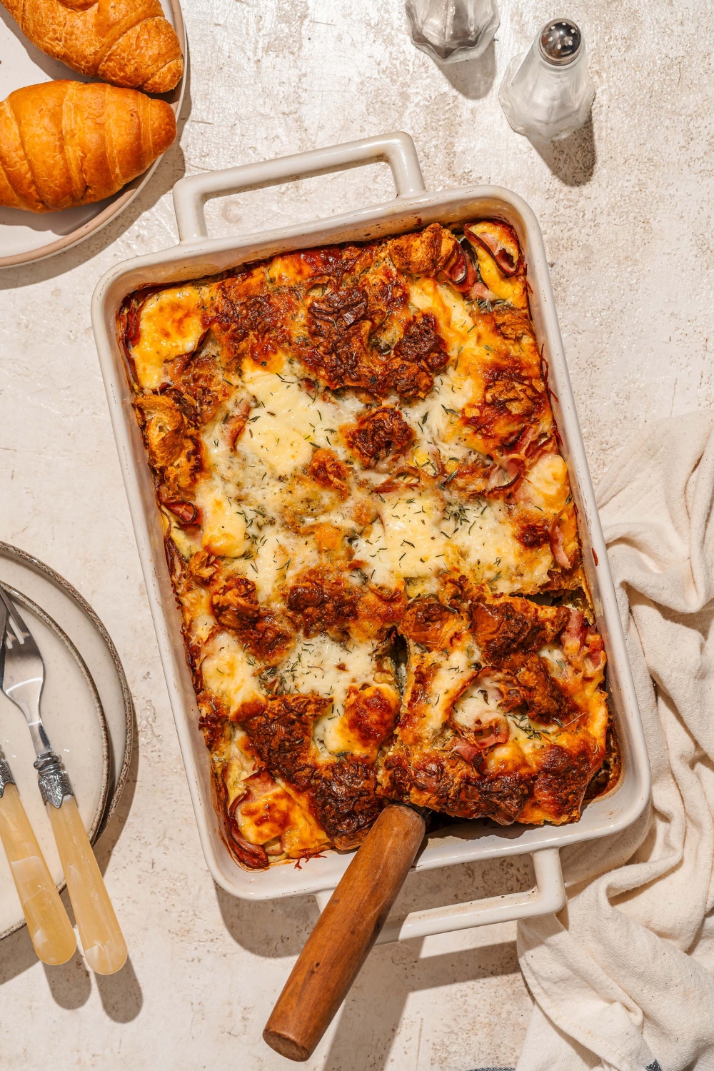 A baking dish with croissant breakfast casserole with ham and cheese. A spatula is inserted in the casserole. The dish is on a tan counter with a white cloth napkin and plate of croissants.