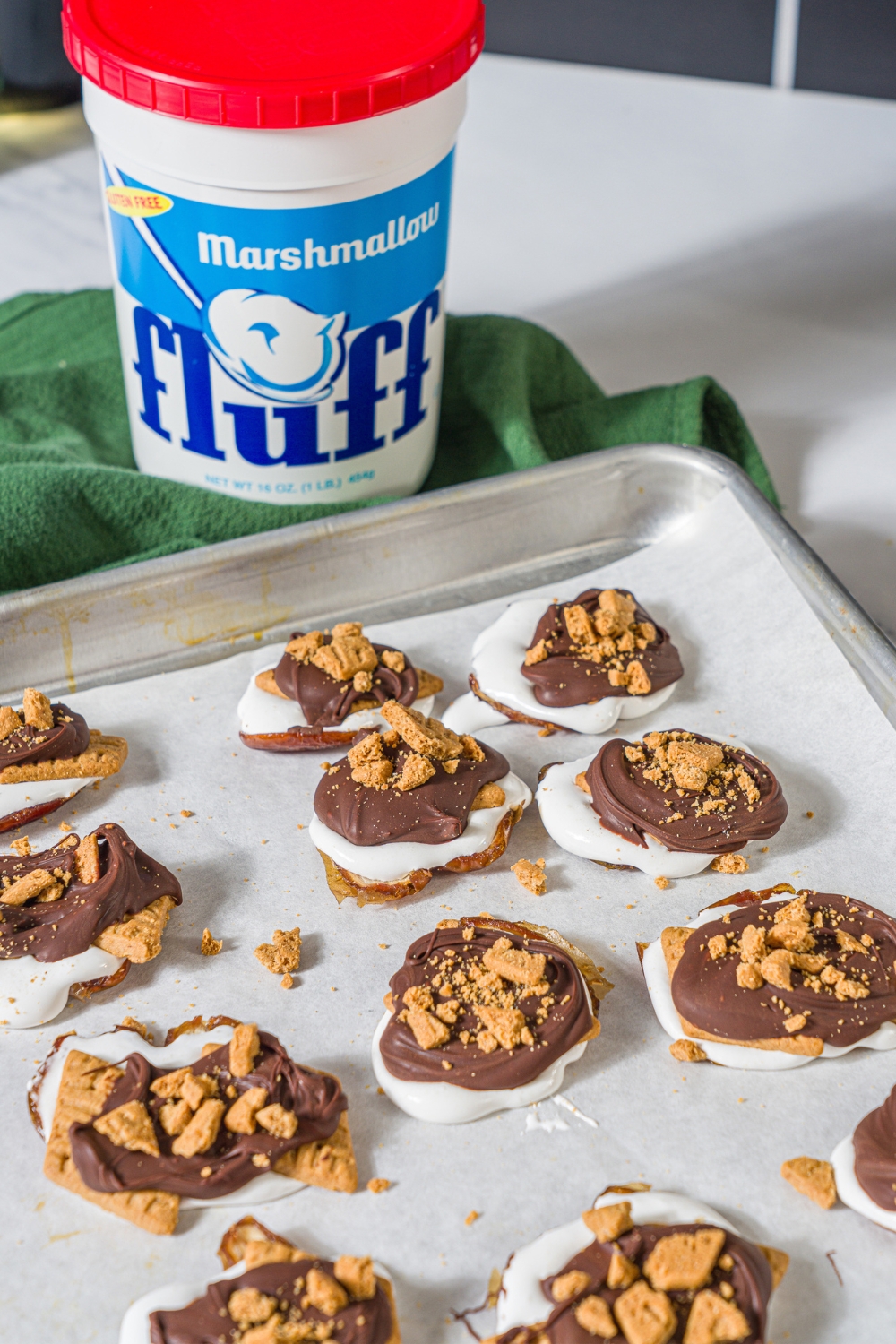 Three rows of three date s'mores on a sheet parchment paper on a baking tray. Behind the tray is a top of marshmallow fluff.