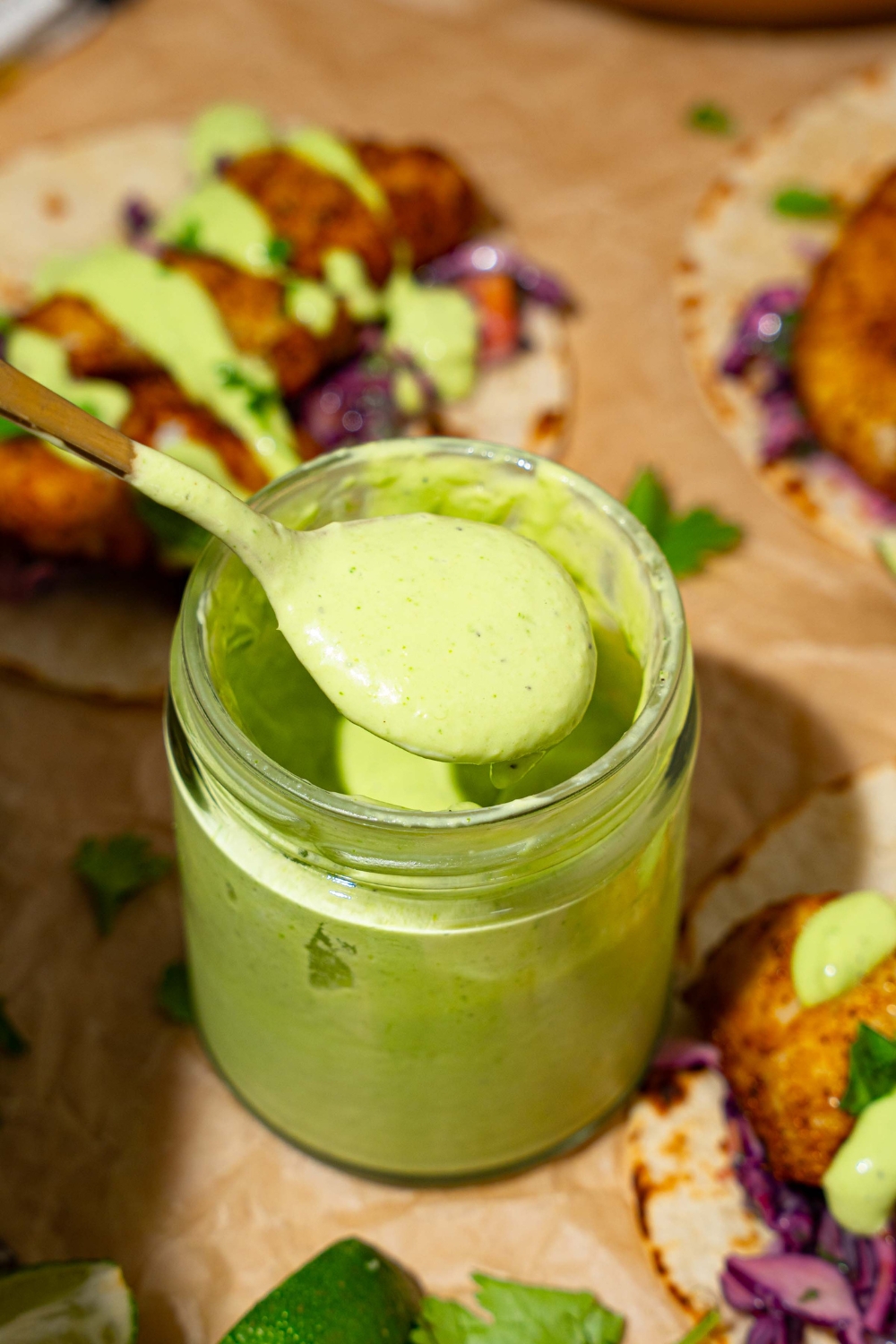 A jar of fish taco sauce on a tan counter lined with parchment paper with several open fish tacos. A spoon is dipping in the sauce.
