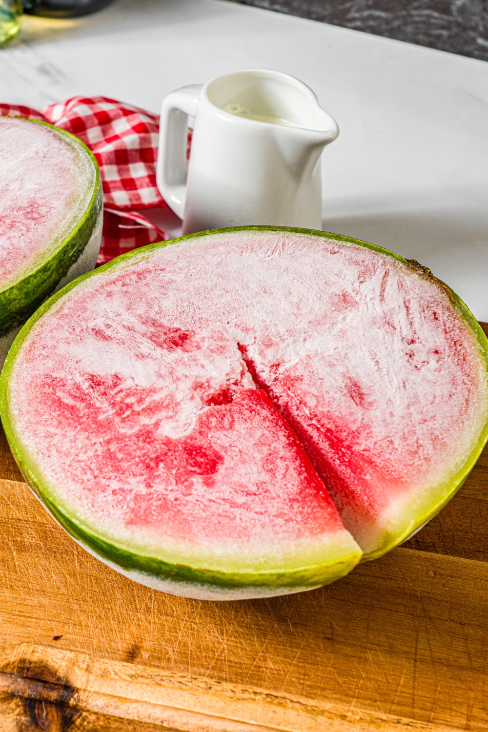 Half of a frozen watermelon on a wooden cutting board with a white pitcher of heavy cream behind it.