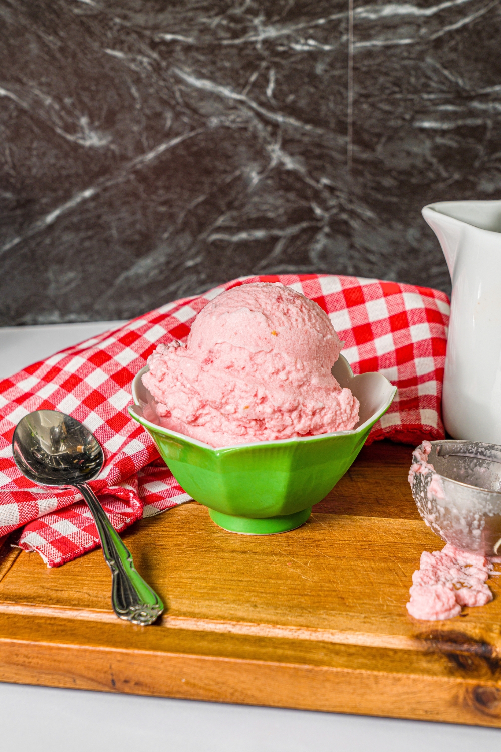 The viral watermelon ice cream in a green bowl that is on a wooden cutting board with a spoon and ice cream scooper next to it.