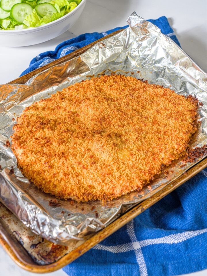A giant crispy ground chicken cutlet on a sheet of aluminum foil on a baking sheet.