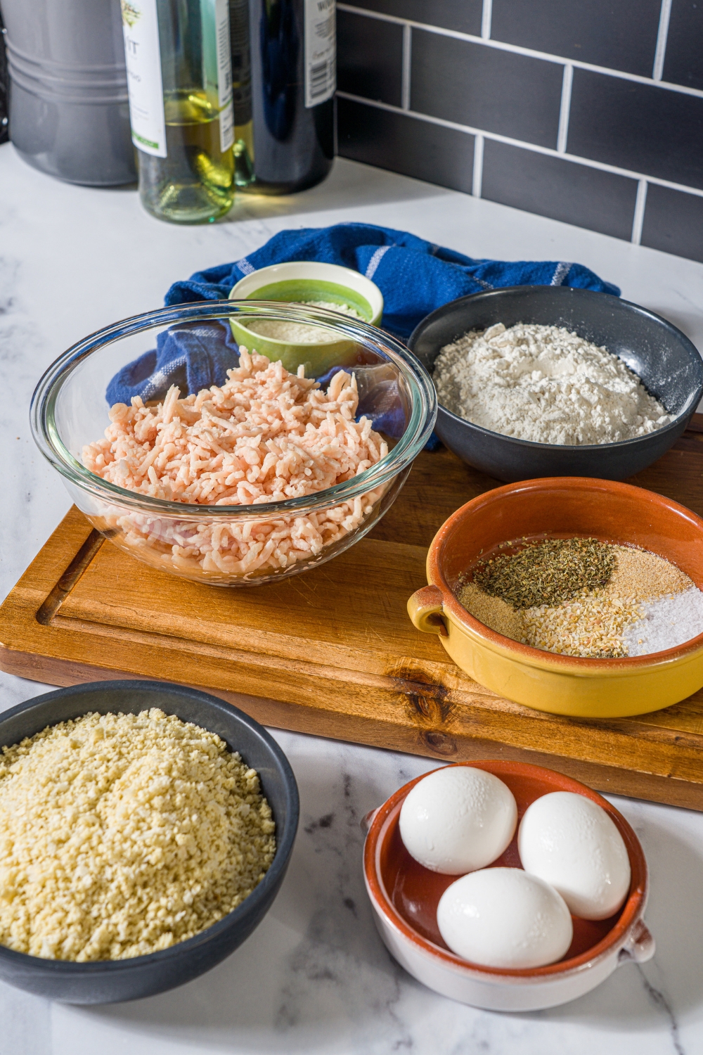 A bowl of ground chicken, a bowl of flour, a bowl of seasonings, a bowl of Panko, breadcrumbs, and a bowl of three eggs.