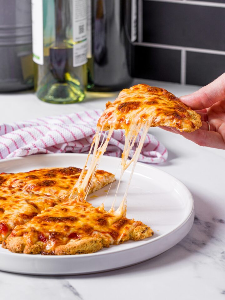 A hand holding a slice of protein pizza above a white plate that has three more slices of the pizza on it.