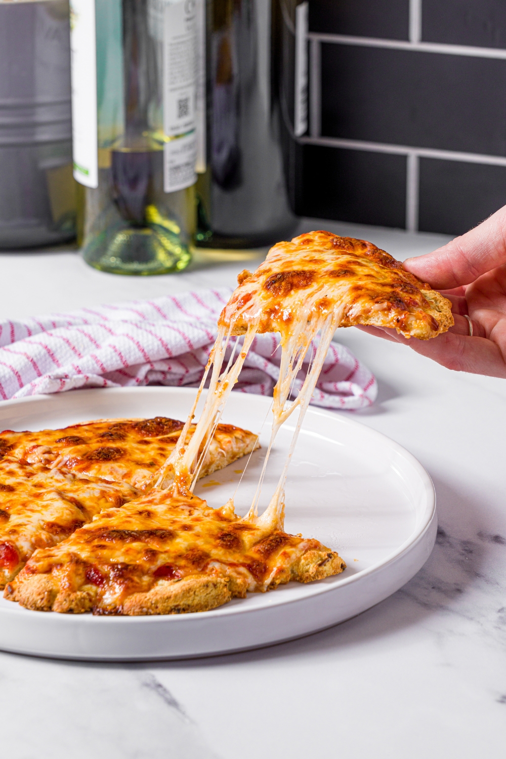 A hand holding a slice of protein pizza above a white plate that has three more slices of the pizza on it.