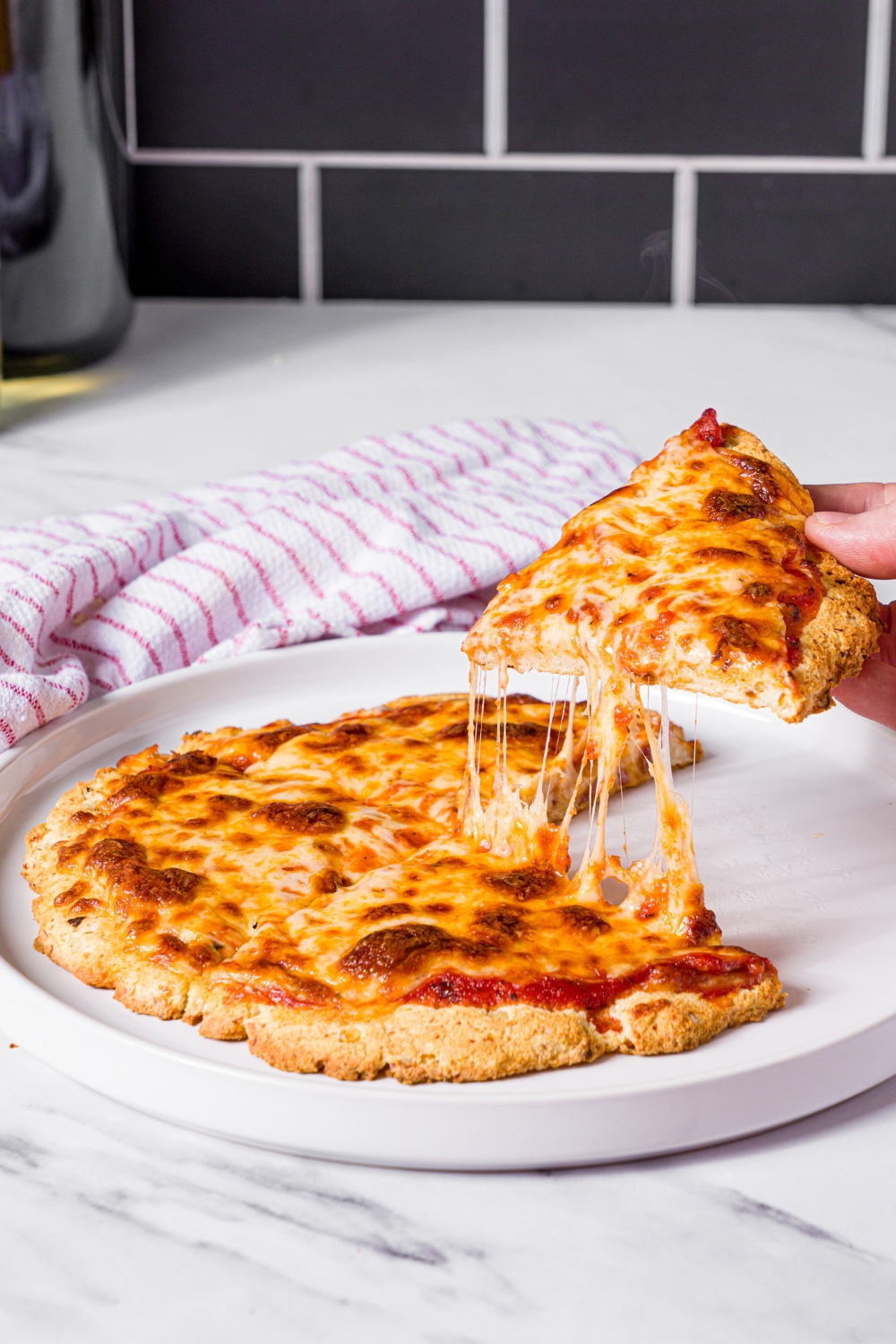 A hand pulling a slice of high protein pizza away from the rest of the pizza on a white plate.