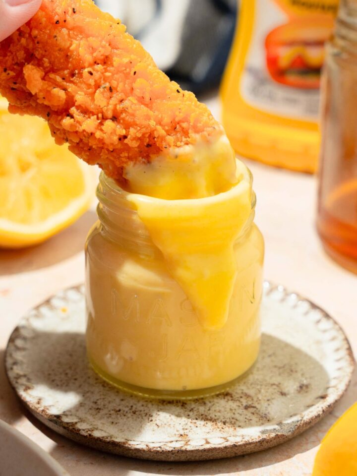 A hand holding a chicken finger that is partially submerged in a glass jar that is filled with a honey mustard, dipping sauce.