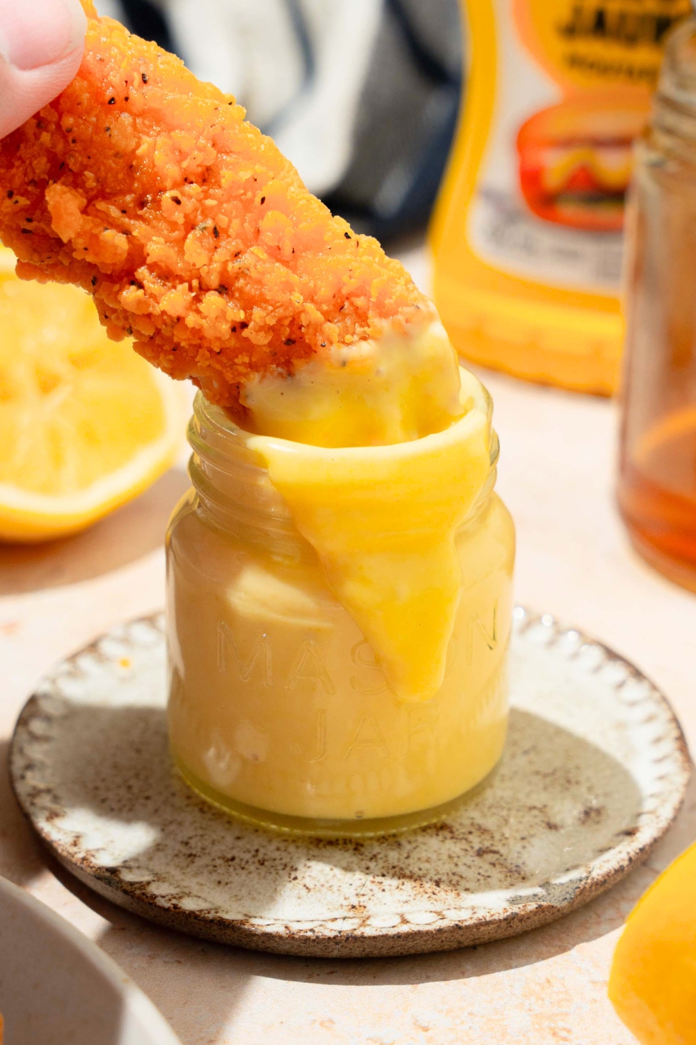 A hand holding a chicken finger that is partially submerged in a glass jar that is filled with a honey mustard, dipping sauce.