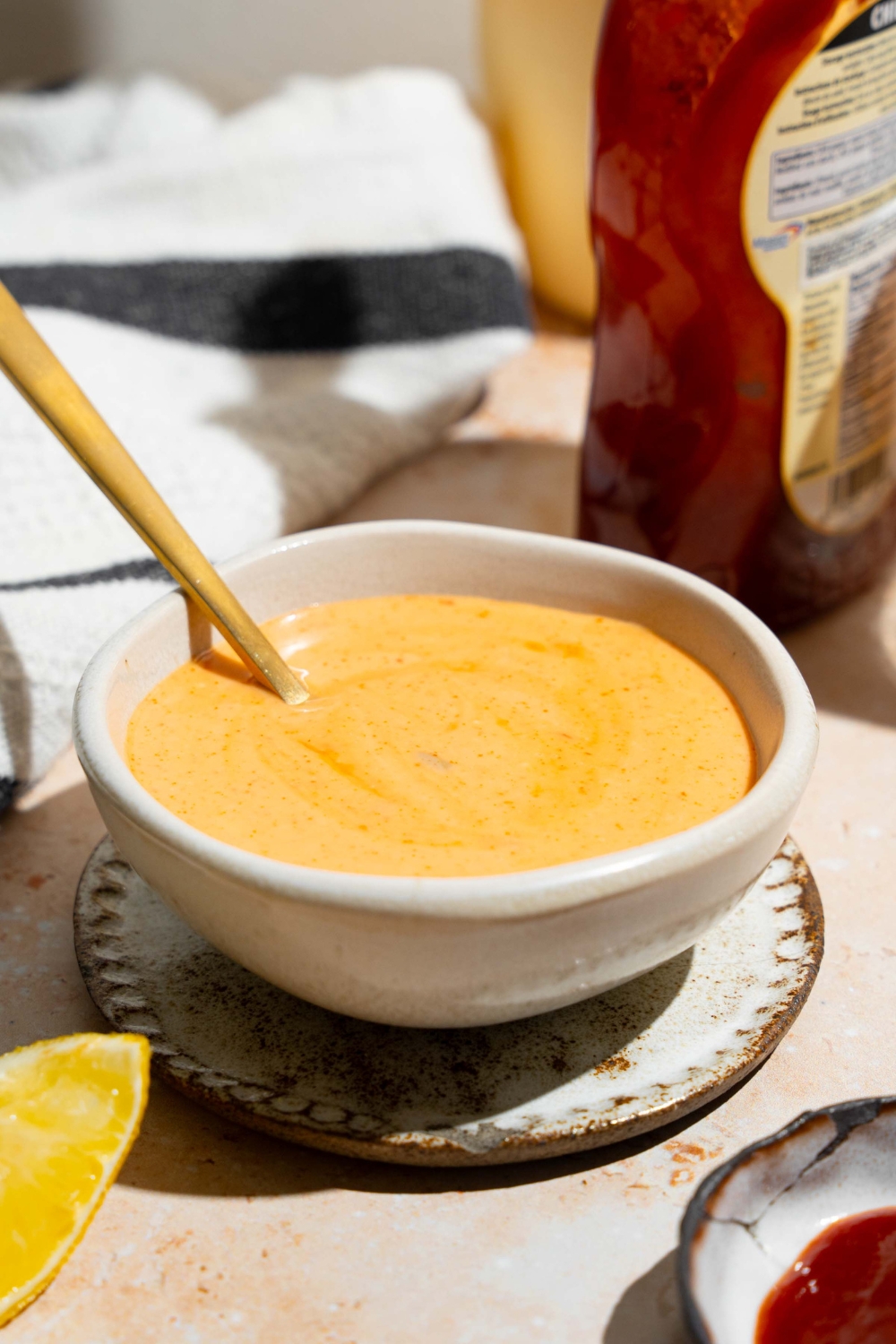 A bowl of honey sriracha sauce with a spoon in the bowl. The bowl is on a tan counter with a jar of honey and bowl of sriracha.