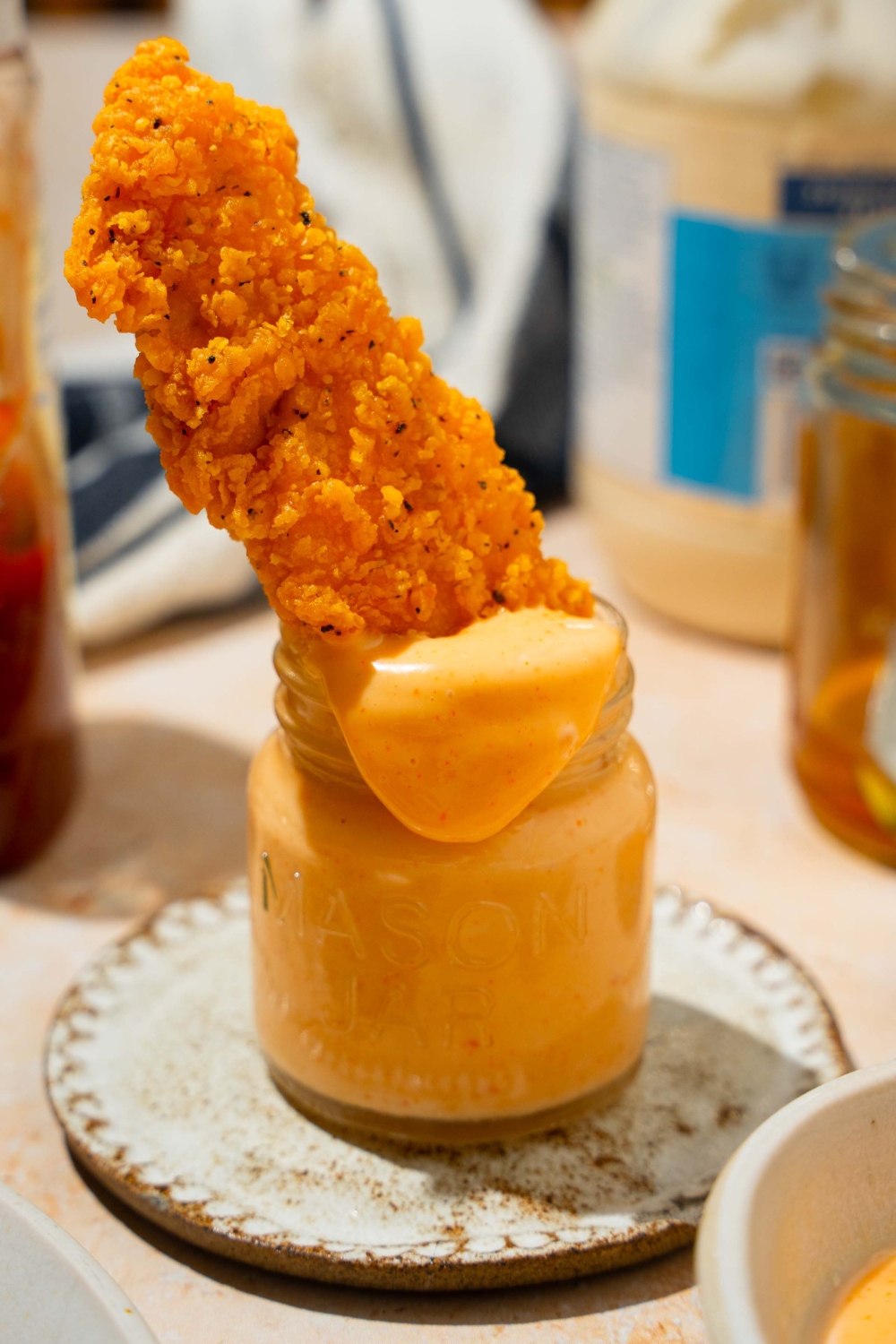 A jar of honey sriracha sauce with a chicken tender dipped in the jar. The jar is on a saucer on a tan counter.