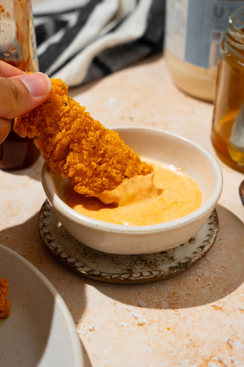 A bowl of honey sriracha sauce with a chicken tender being dipped into the bowl. The bowl is on a tan counter with a jar of honey.