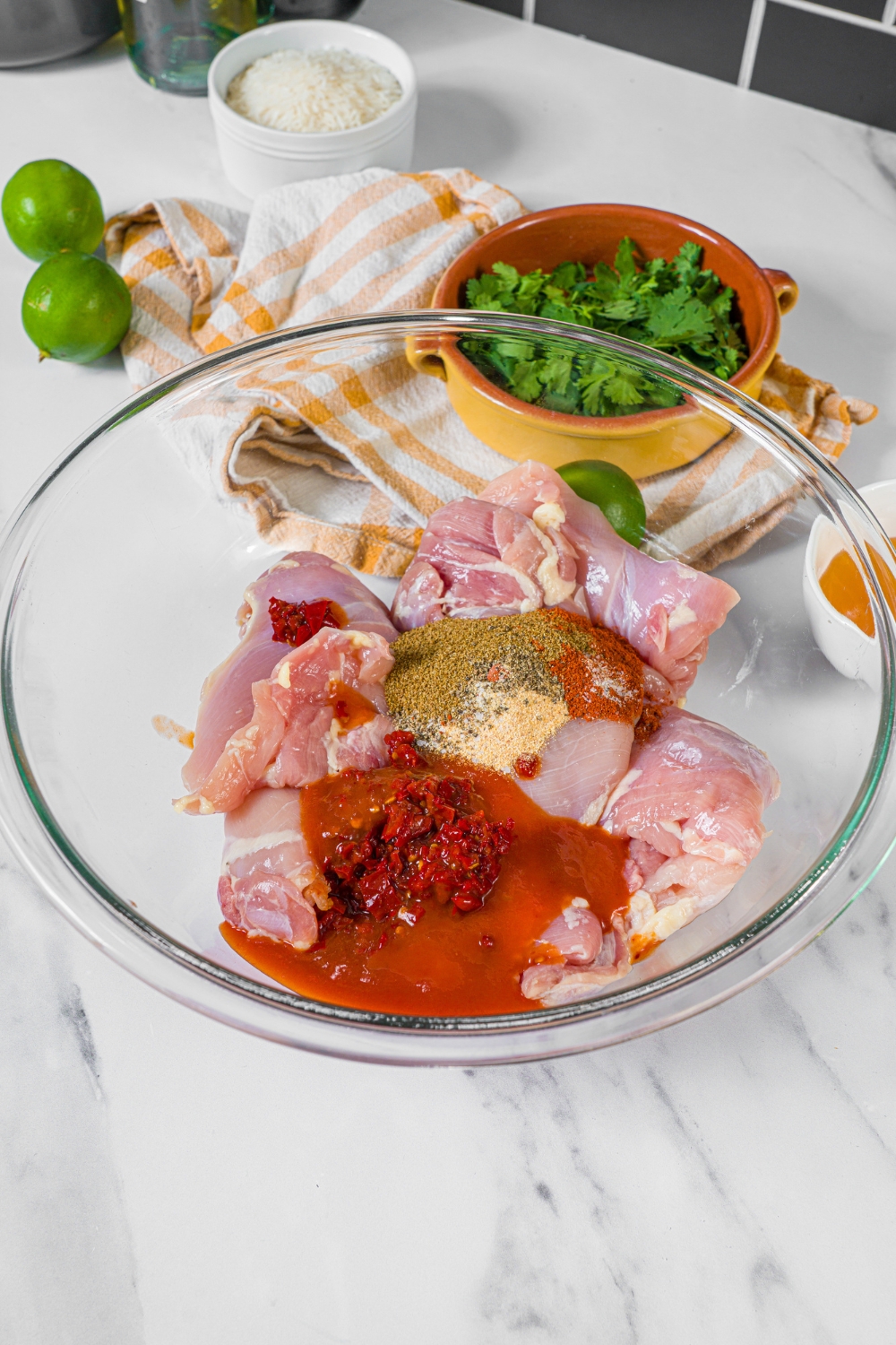 A glass bowl of chicken thighs and a bunch of seasonings on them in a glass bowl.