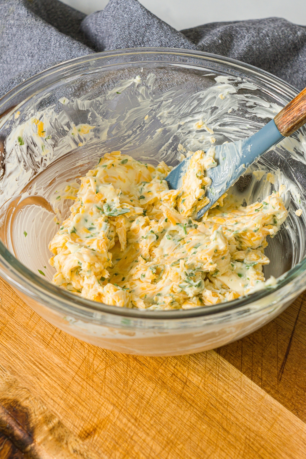 A glass bowl with a spatula mixing cheesy garlic mixture. The bowl is on a wooden counter.