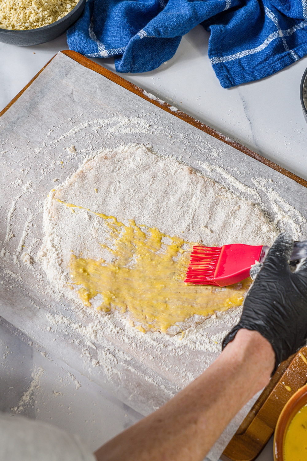 Red brush brushing a giant ground chicken cutlet mixture with egg wash. The ground chicken mixture is on a flowered sheet parchment paper on a baking sheet.
