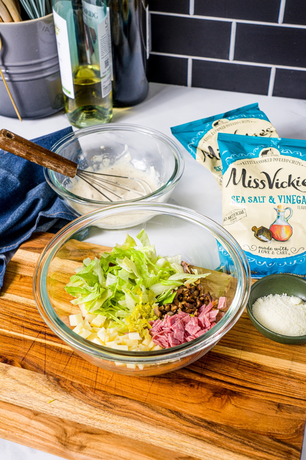 Chopped lettuce, chopped salami, chopped, provolone cheese, and chopped black olives in a glass bowl on a wooden cutting board. Behind that are two salt and vinegar, chip bags, and a bowl of Italian creamy dressing.