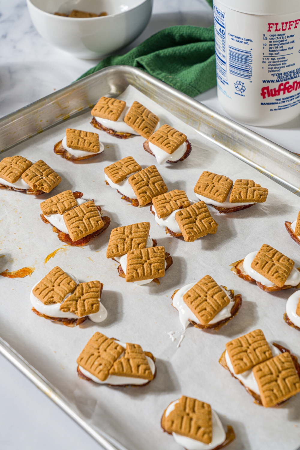 A parchment line baking sheet that has a bunch of smashed dates with marshmallow, fluff, and graham crackers on top of it.
