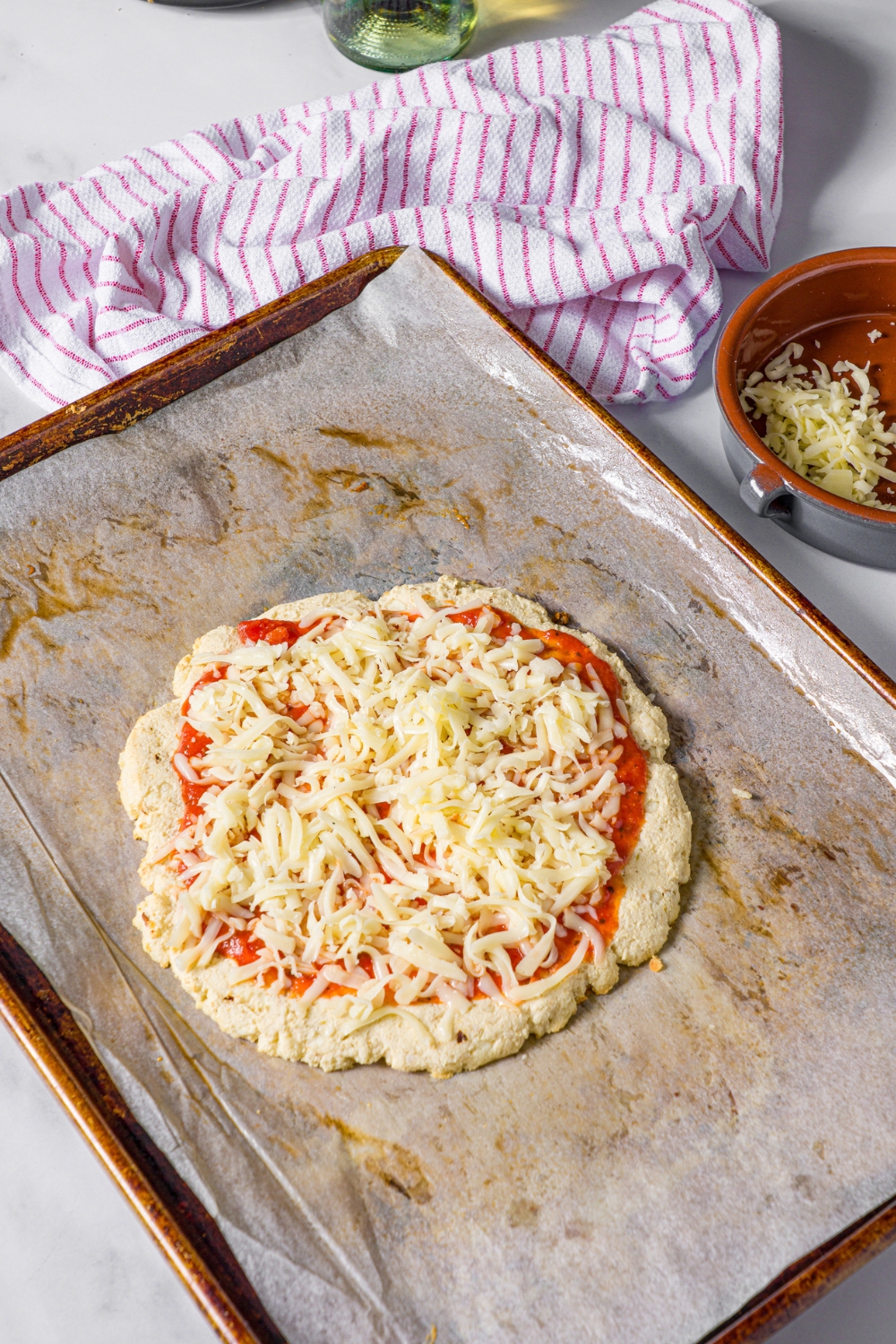 Shredded mozzarella cheese on top of marinara sauce on a protein pizza crust on a sheet of parchment paper that's on a baking tray.
