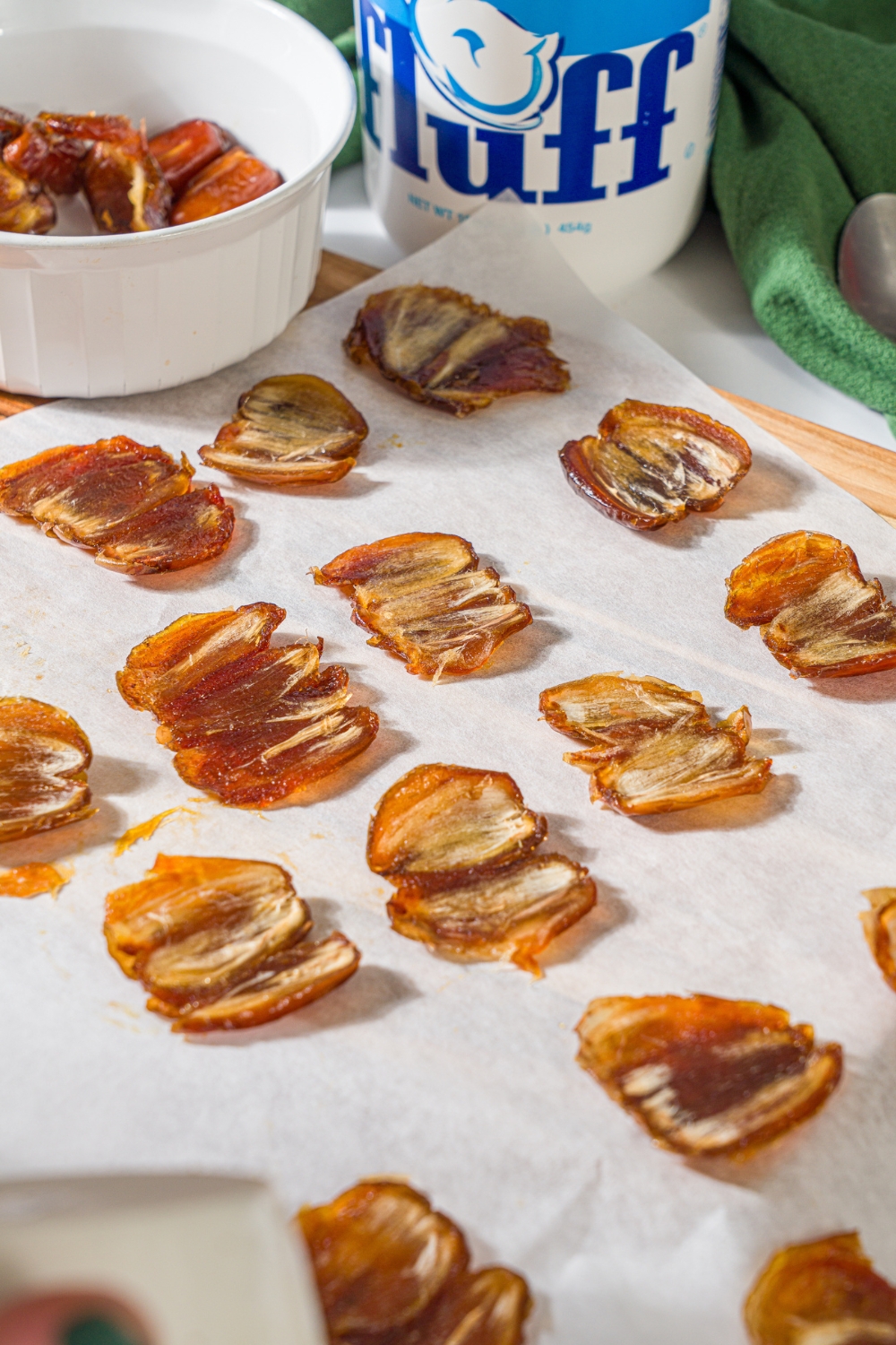A bunch of smashed dates on a sheet of parchment paper with a bowl of dates and a tub of marshmallow fluff behind it.