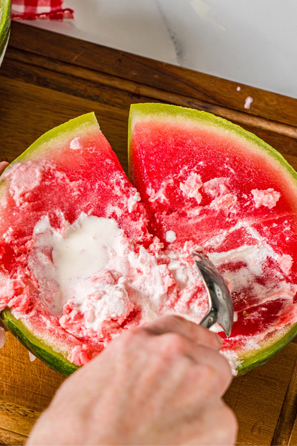 A hand holding a spoon submerged in half of a frozen watermelon that has heavy cream in it.