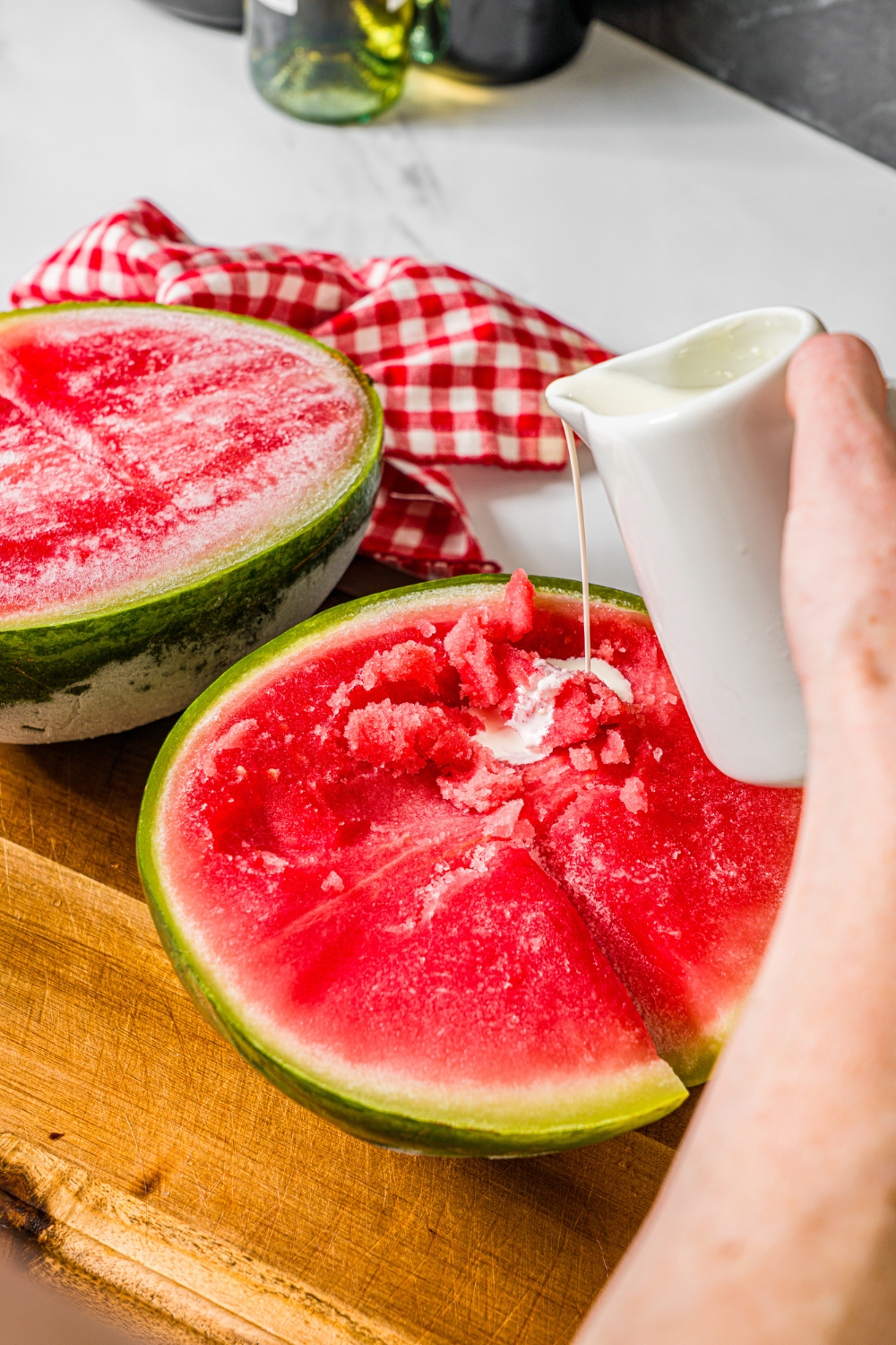 A hand pouring heavy cream from a white pitcher into a half of a watermelon that is on a wooden cutting board. Another half of a frozen watermelon is behind it.