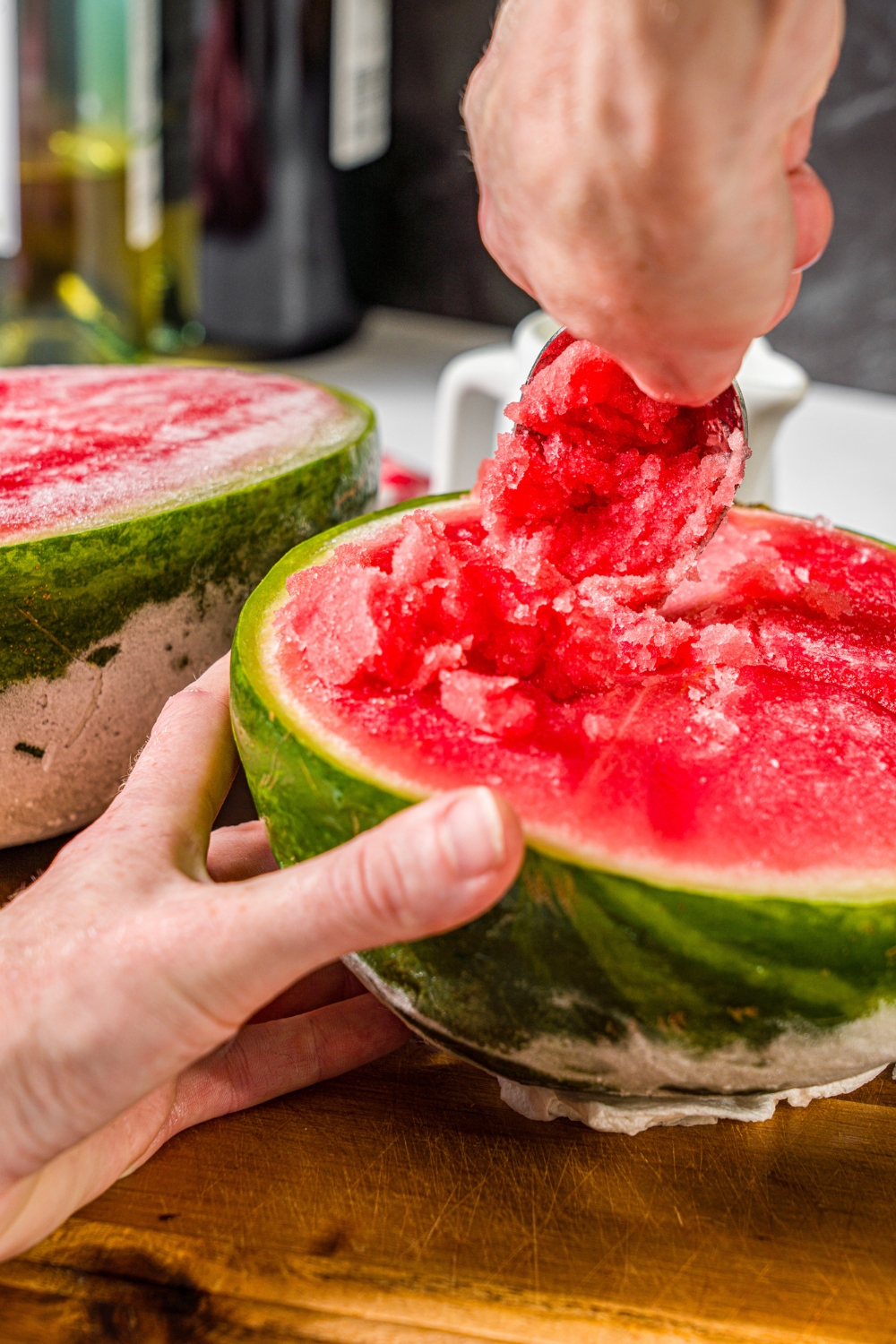 A hand holding a spoon scooping out the inside of a half watermelon with the other hand holding the watermelon.