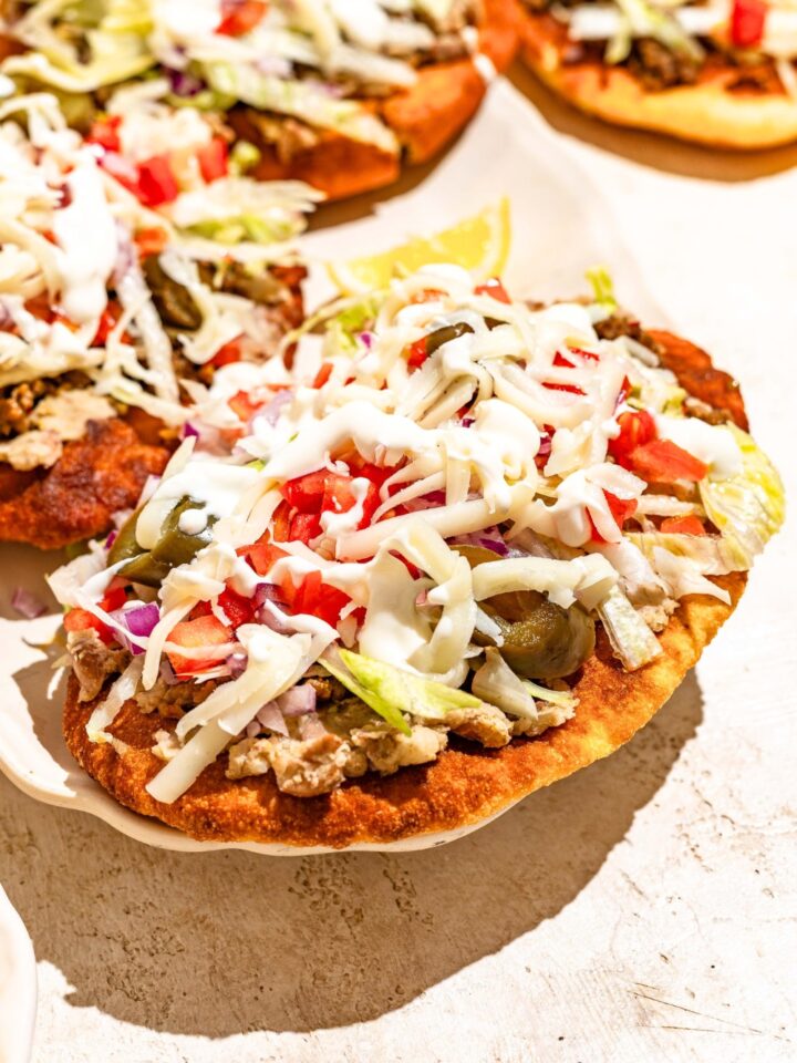 An Indian taco that has ground beef, shredded cheese, jalapenos, tomatoes, lettuce, onion, and a white sauce on top of Indian fry bread.